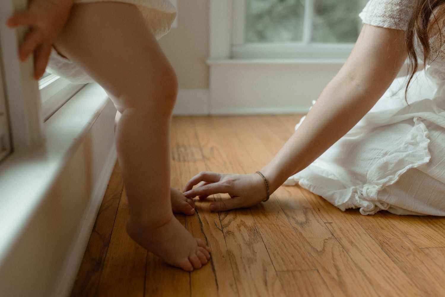 A woman in a white dress kneels on hardwood floor, lovingly touching a toddler's bare feet by a window.