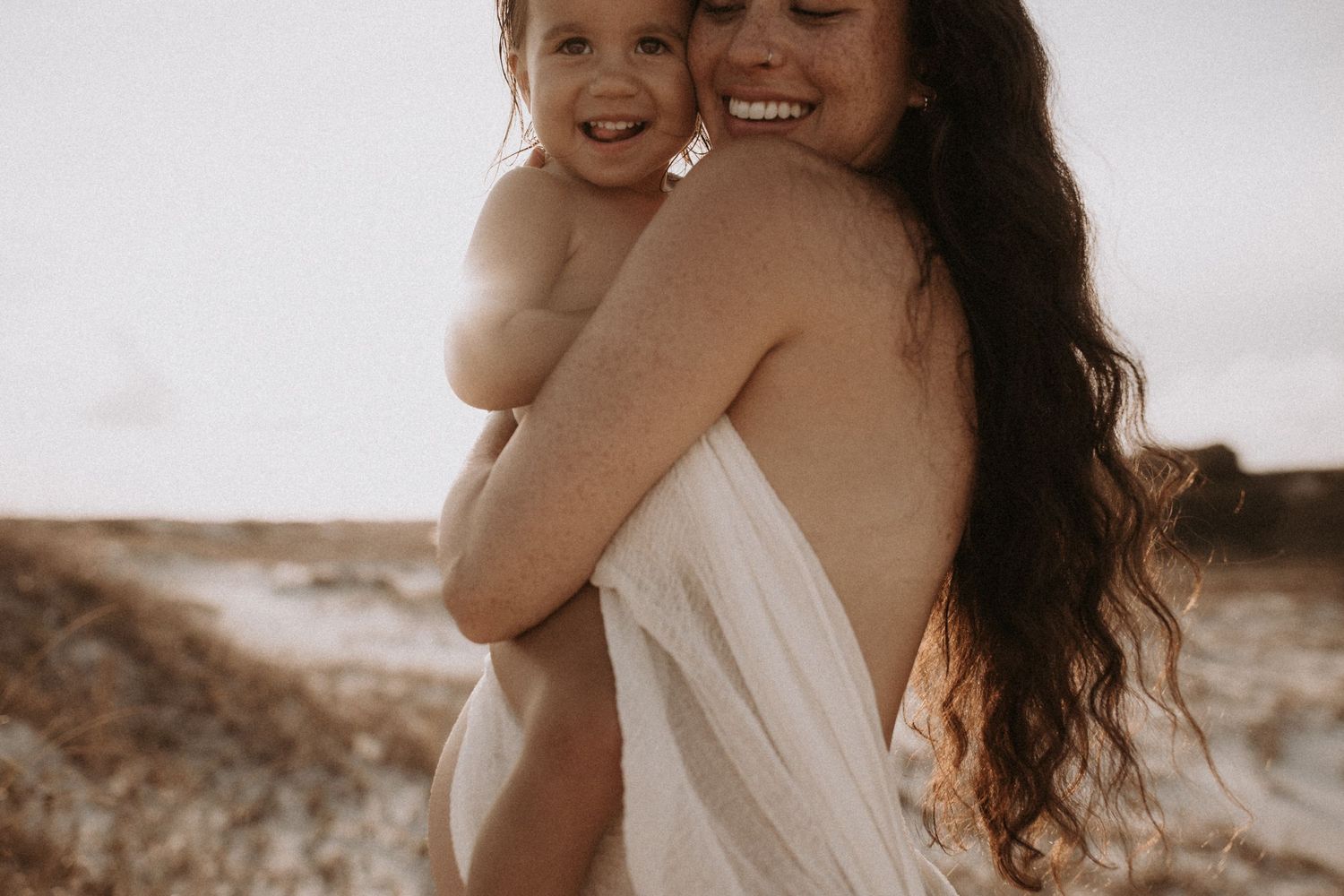 Smiling mother with long dark hair holds laughing toddler on sandy beach wrapped in white fabric.