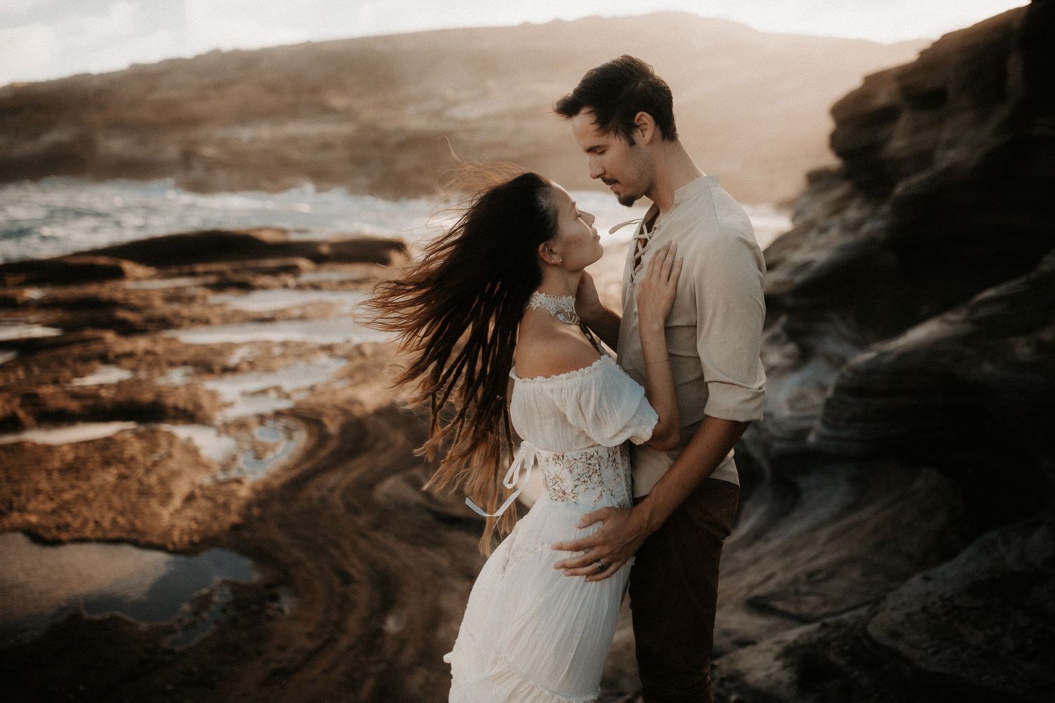 Couple embracing romantically on rocky coastal cliffs at golden sunset, woman in white dress with hair blowing in wind.