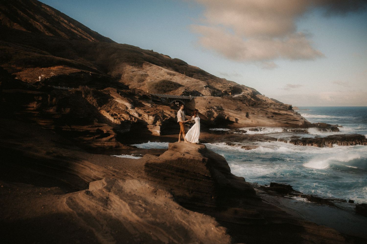 Couple in wedding attire standing on rocky coastal cliffs with ocean waves and dramatic hillside backdrop.