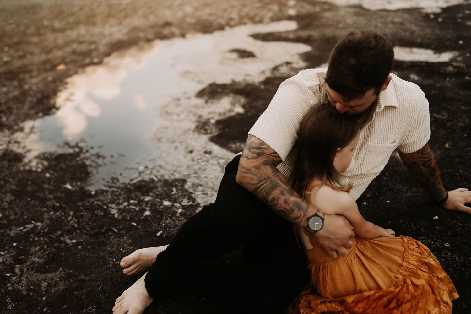 A tattooed man leans over a woman in an orange skirt on dark ground near a reflective puddle.