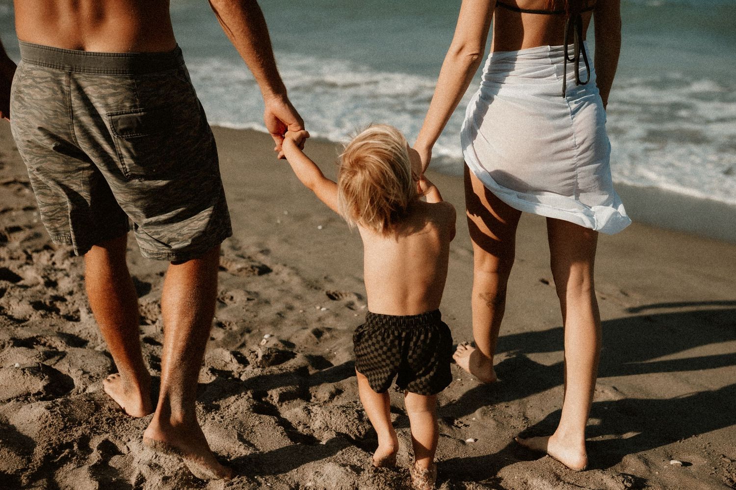Parents holding toddler's hands at ocean's edge, walking barefoot on wet sand together.