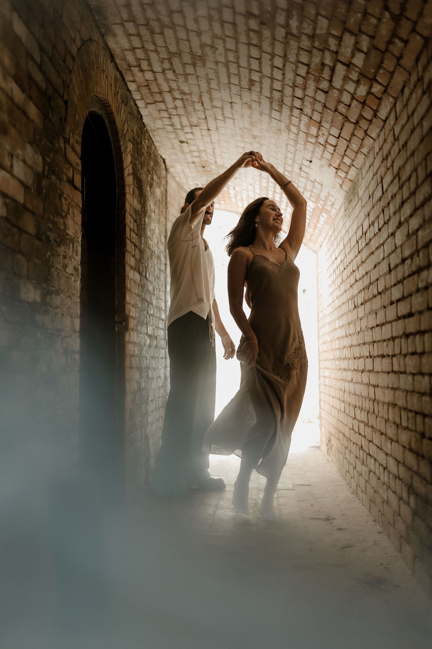 Couple dancing elegantly in a misty brick tunnel corridor with dramatic backlight creating a romantic atmosphere.