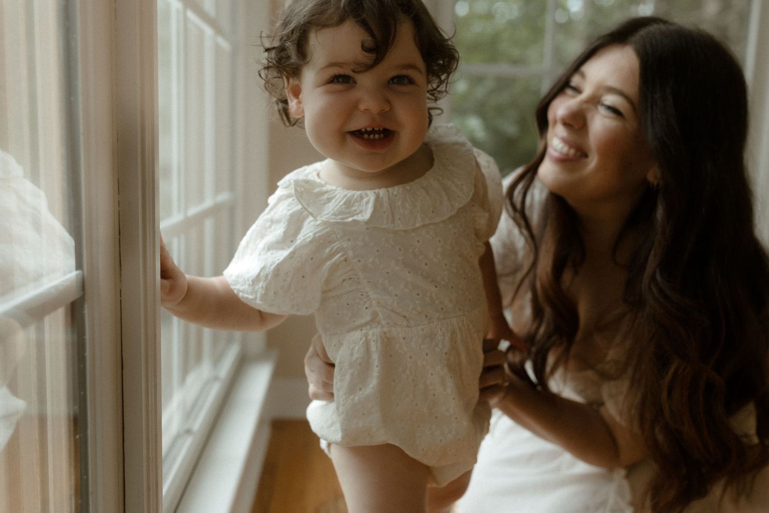 A smiling mother holds her happy toddler girl by a bright window indoors.