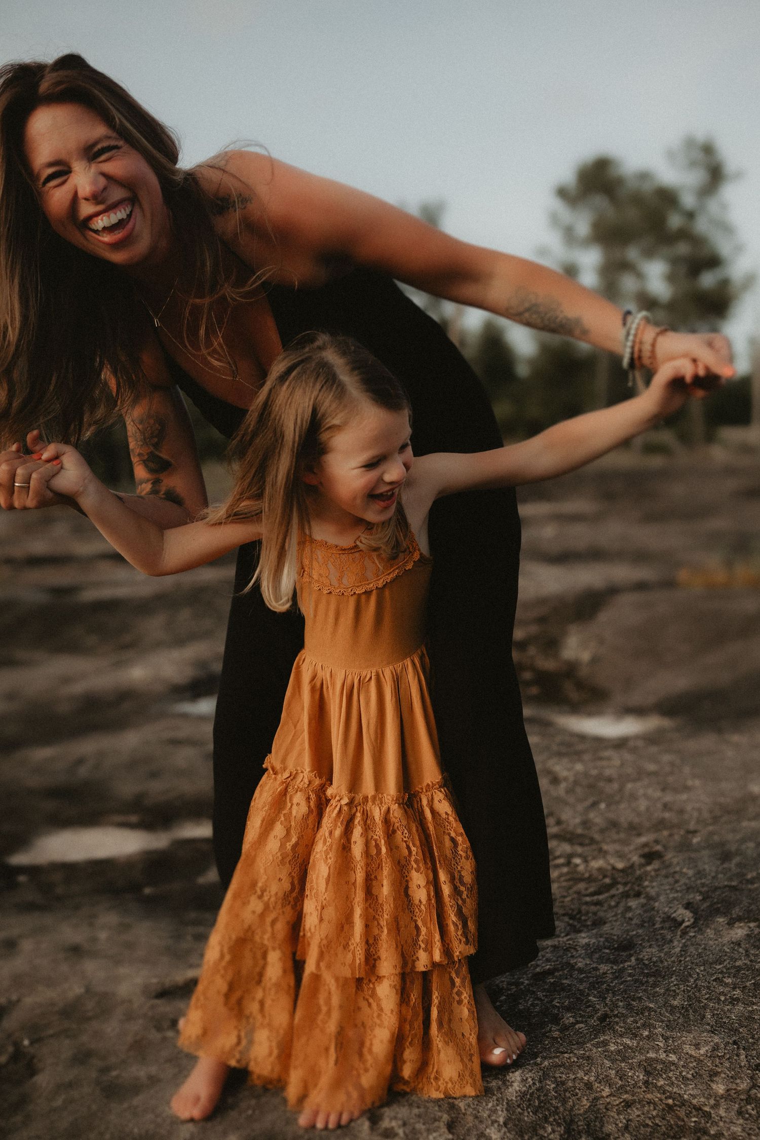 A laughing mother in black dress holds hands with her smiling young daughter in an orange tiered dress outdoors.