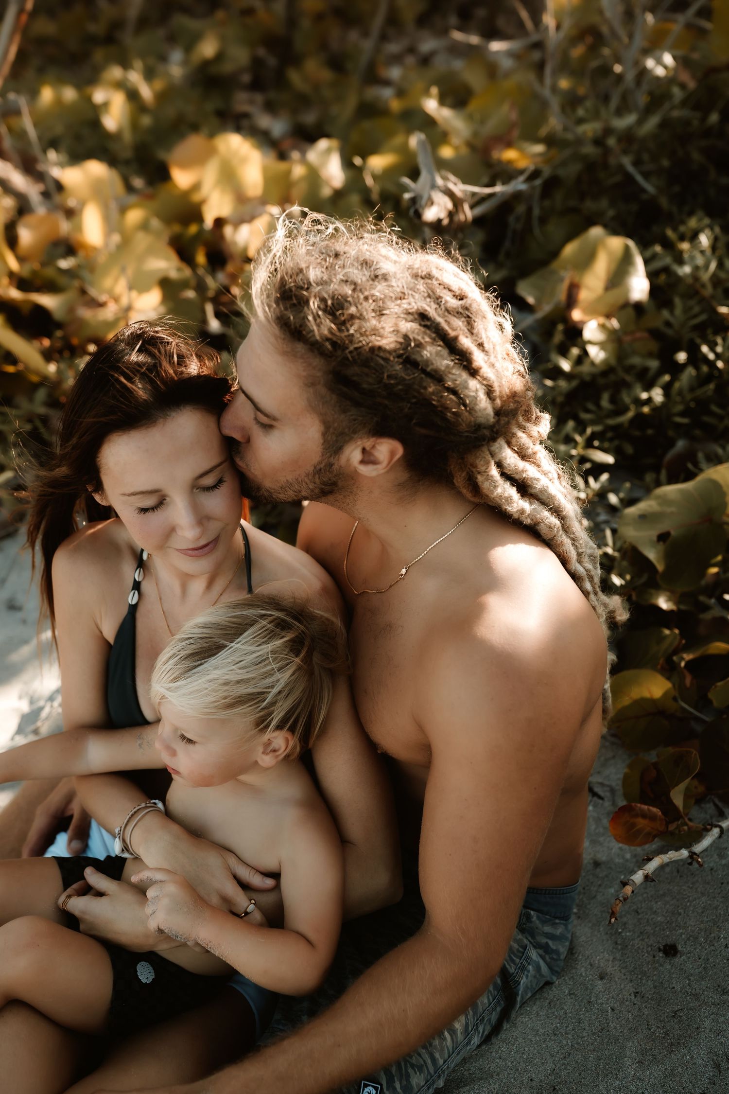 A loving couple with dreadlocks kisses while holding their young blonde child on a sunny beach.