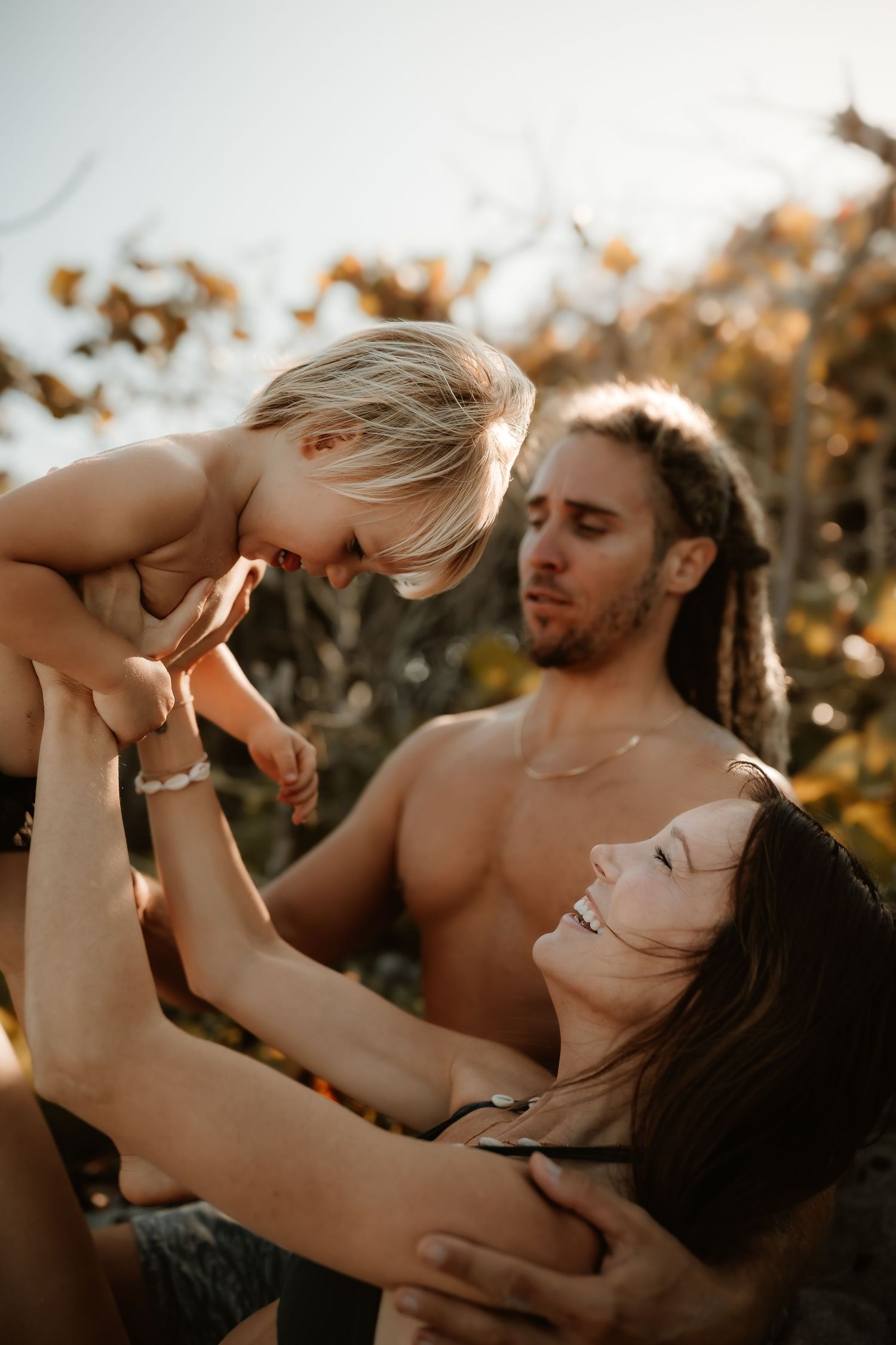 Shirtless man with dreadlocks and woman in bikini joyfully lifting blonde toddler outdoors.