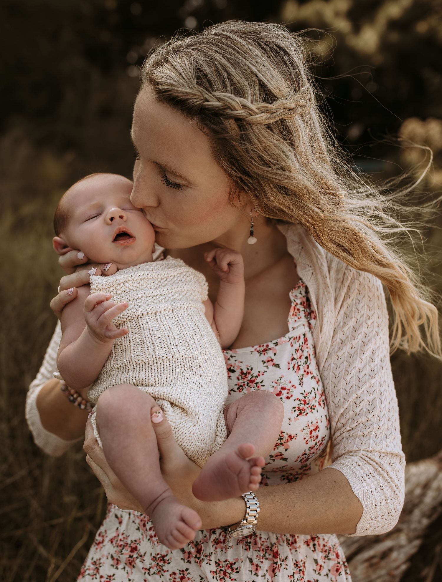 Mother kissing newborn baby wearing cream knit outfit in natural outdoor setting.