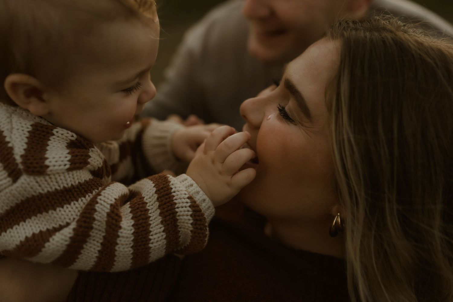 A mother tenderly holds her baby in a brown striped sweater as the child playfully touches her face outdoors.
