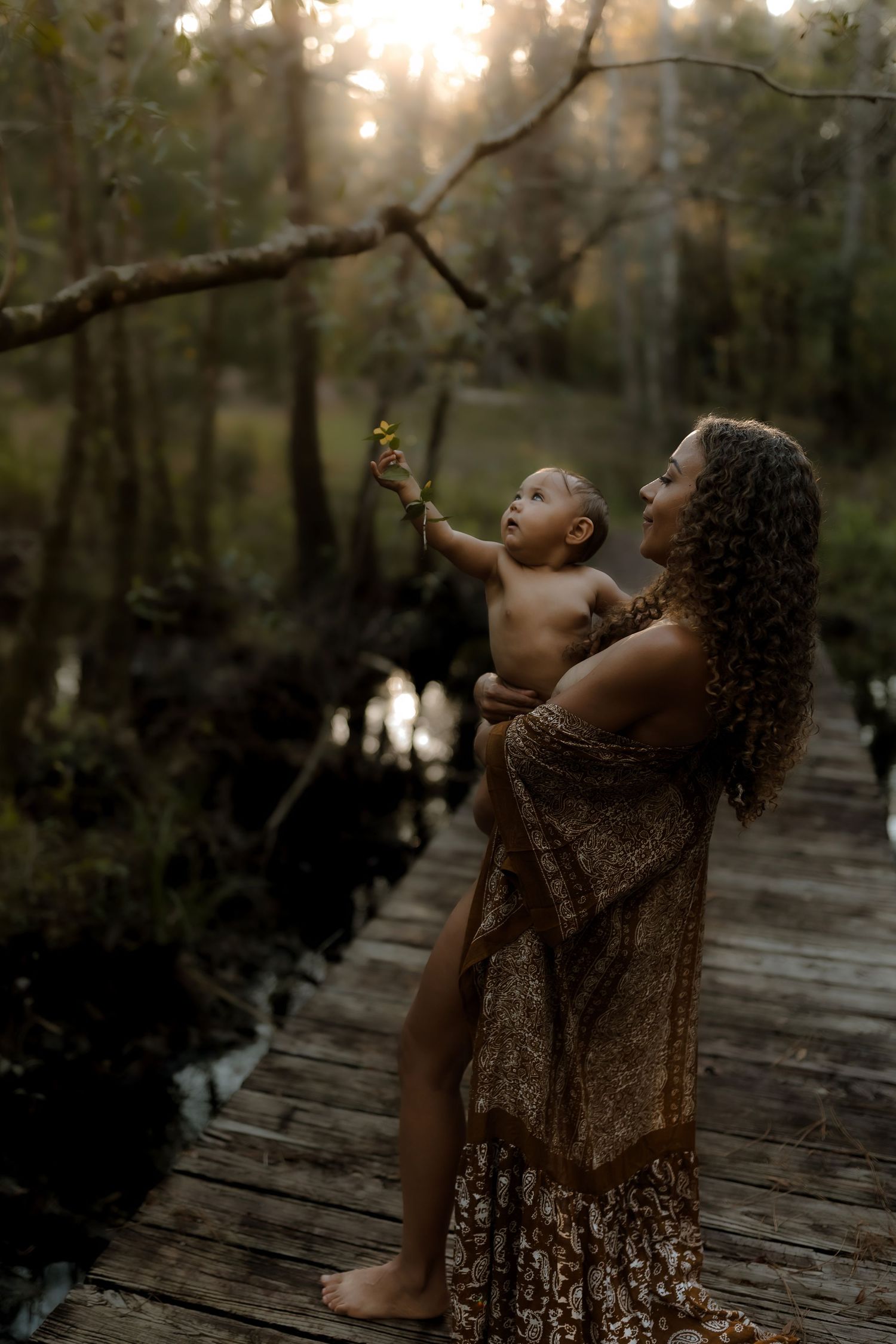A mother holds her baby up toward a tree branch on a wooden bridge in a sunlit forest.