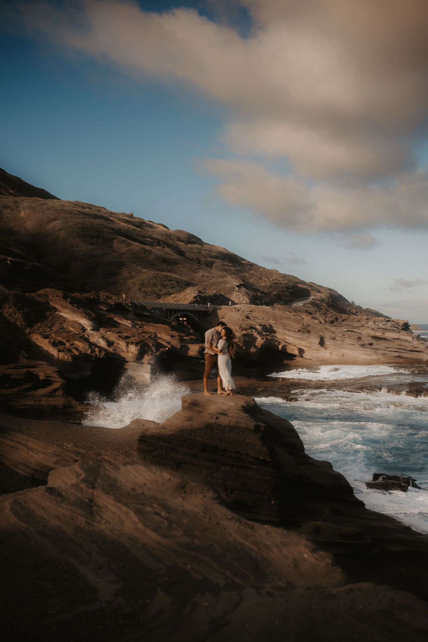 Couple embracing on rocky coastal cliffs with ocean waves crashing below under a dramatic cloudy sky.