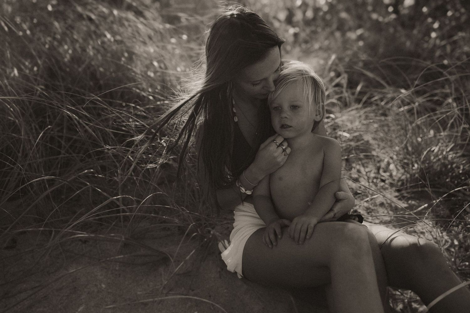 A mother tenderly holds her toddler outdoors among tall grass in a serene black and white nature portrait.