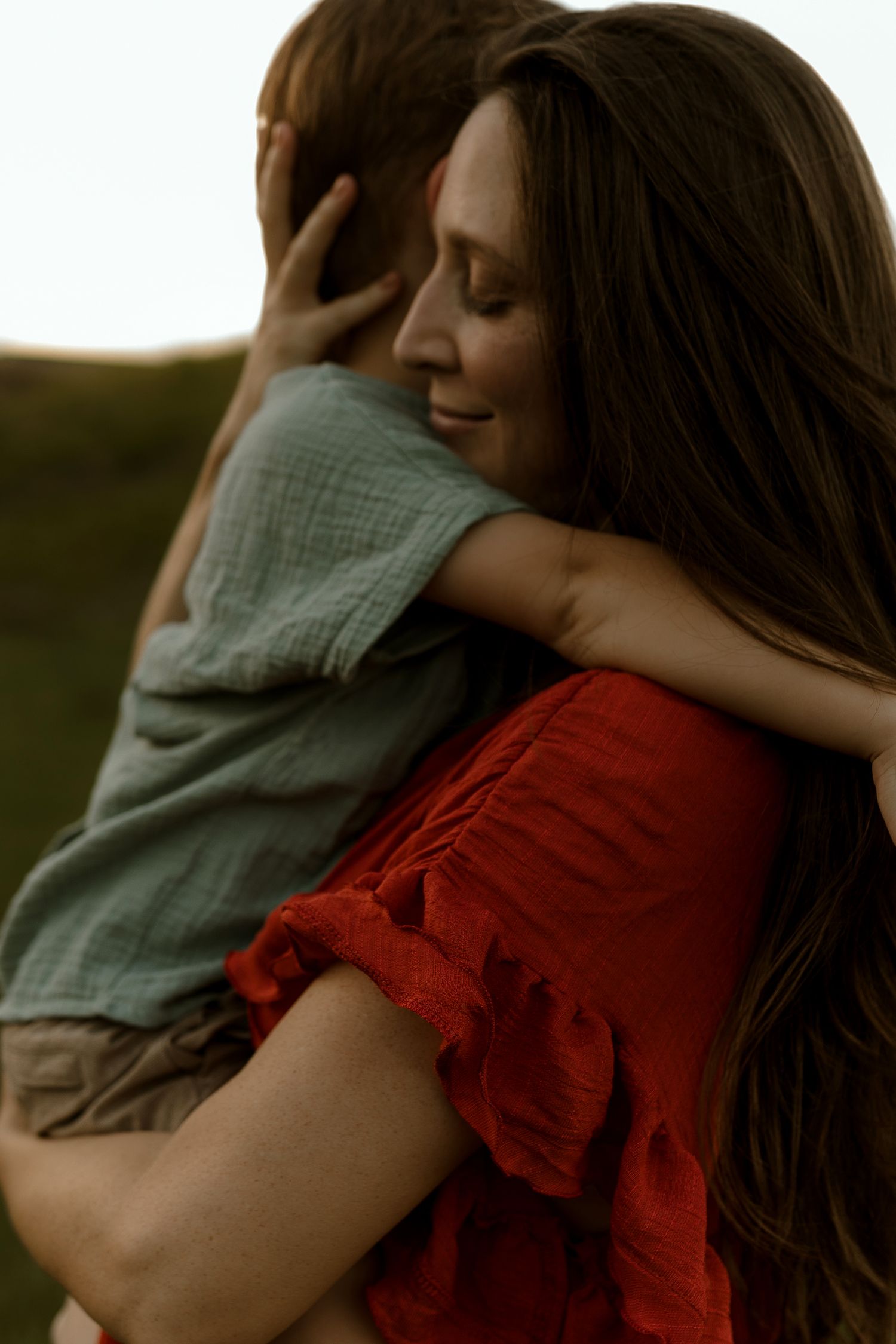 A mother with long brown hair hugs her young son outdoors in a warm red dress at sunset.