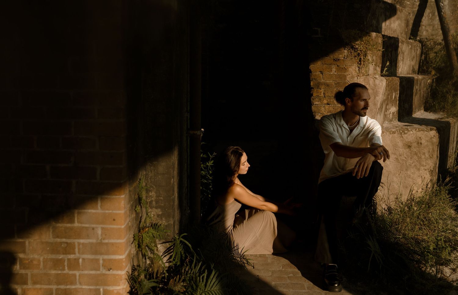 A couple shares an intimate moment in a dimly lit doorway, surrounded by brick walls and lush greenery at night.