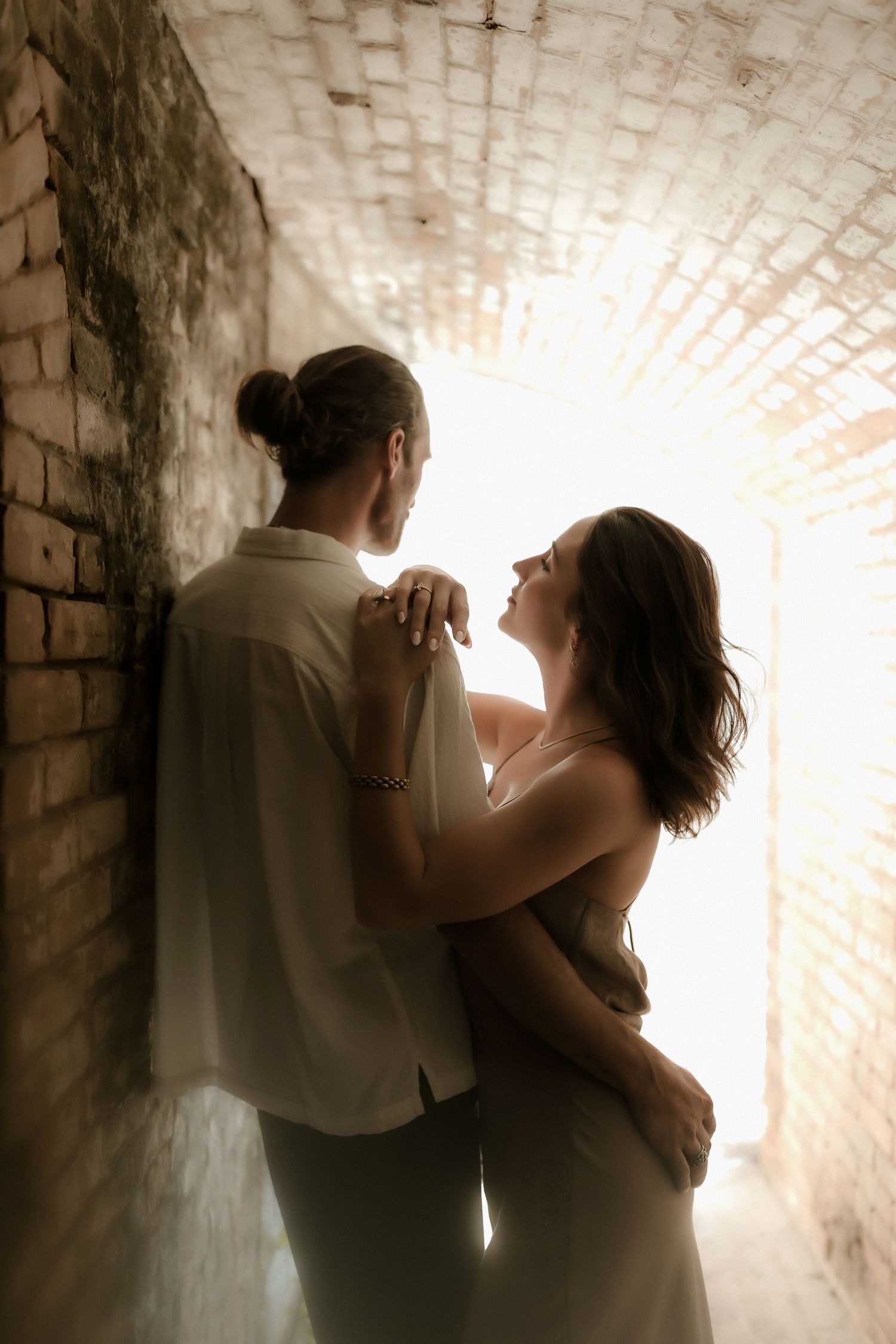 Romantic couple embracing in a sunlit brick tunnel archway, man with bun hairstyle holding woman in white dress.