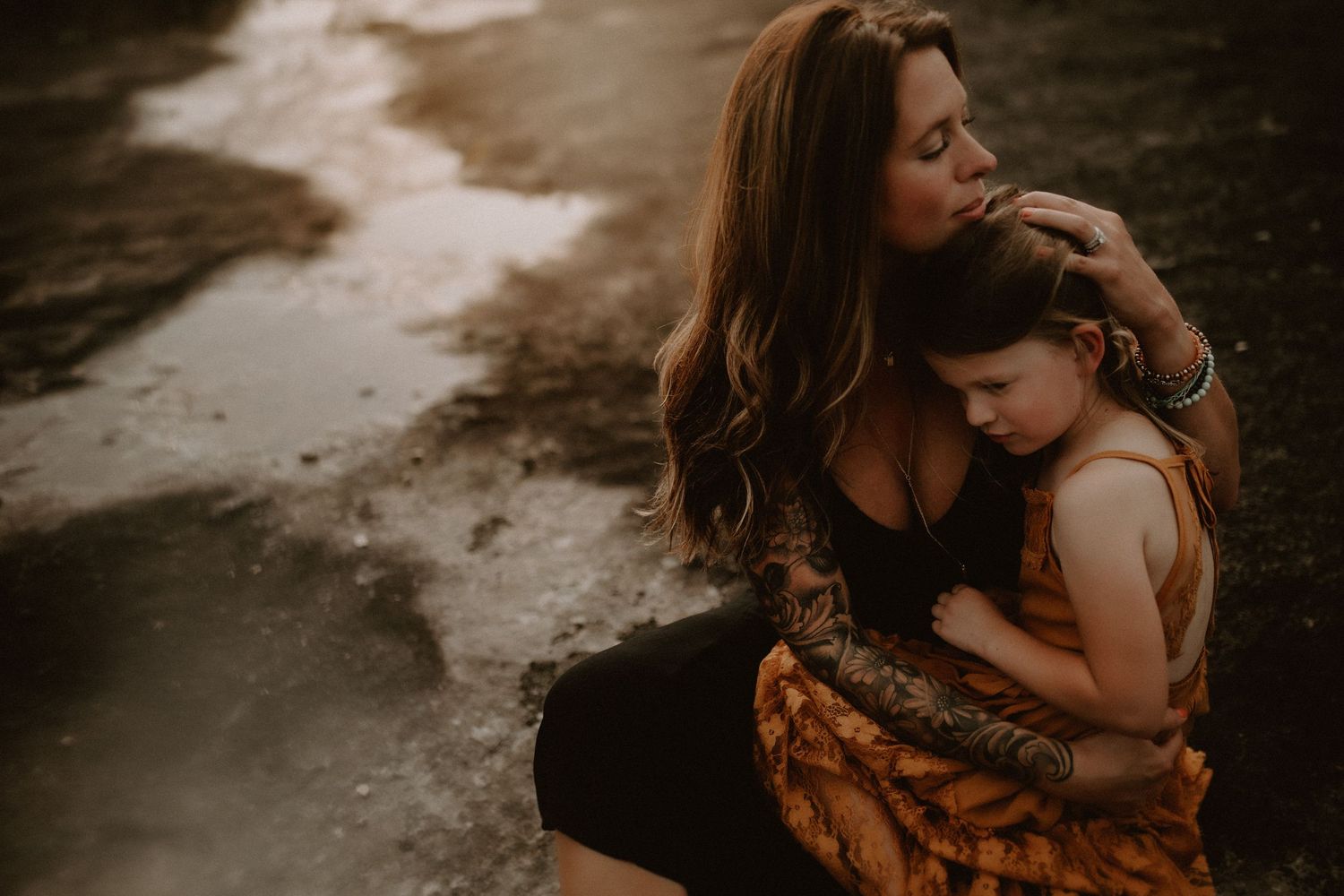 A tattooed mother tenderly embraces her young child outdoors in warm golden light.