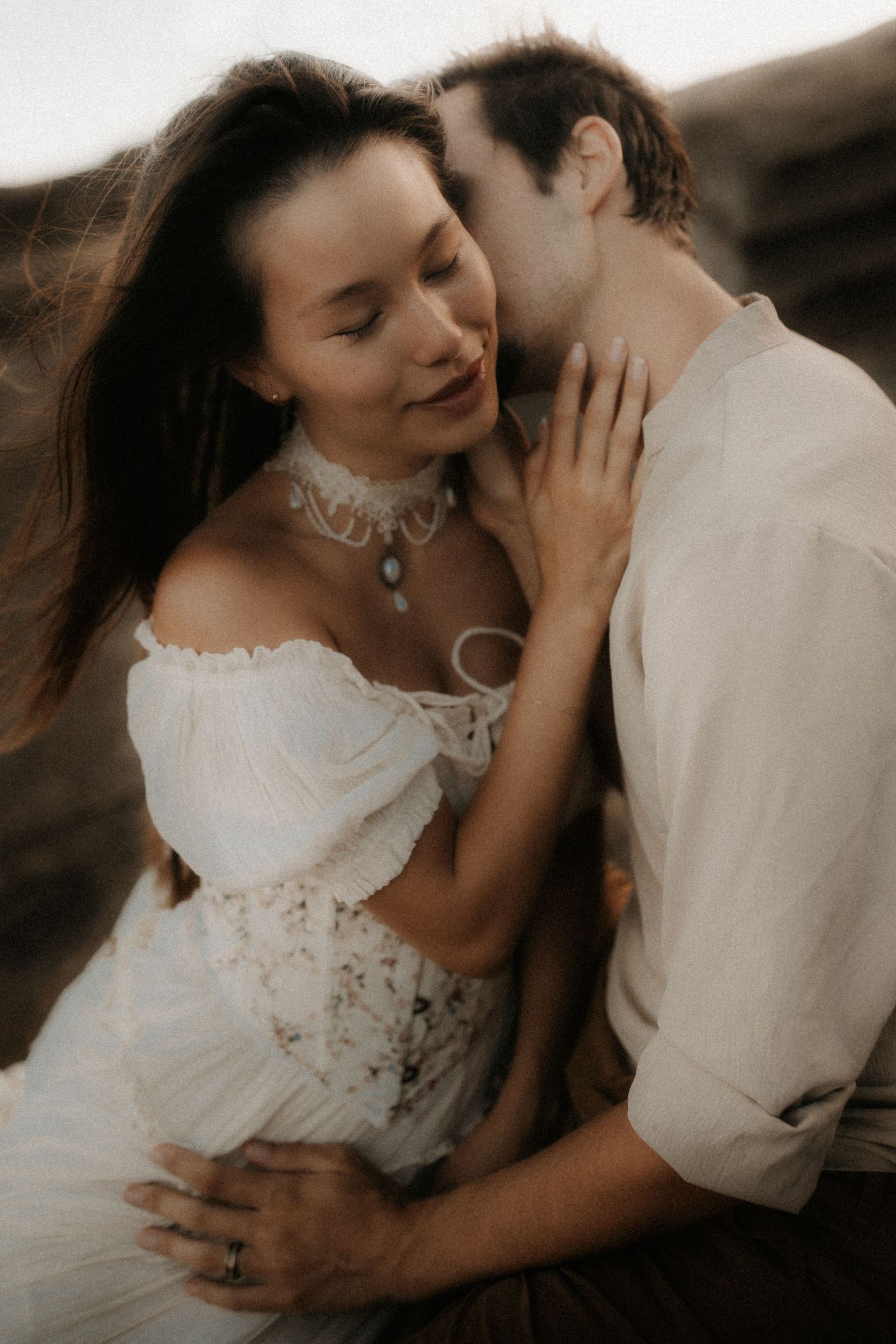 A couple in white bohemian attire share a romantic embrace, the groom kissing the bride's cheek outdoors.