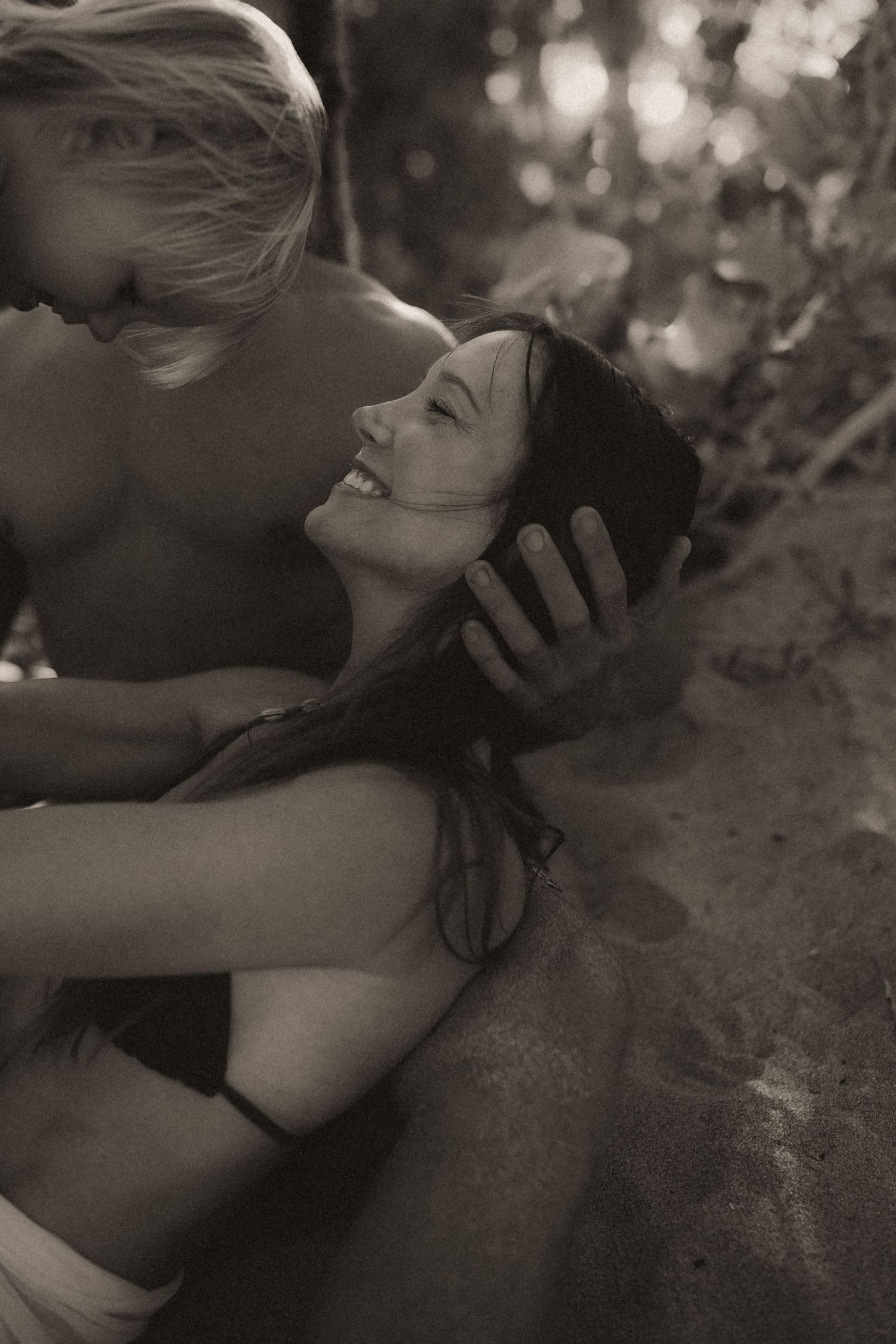 Black and white photo of a smiling woman and shirtless man embracing romantically on a sandy beach.