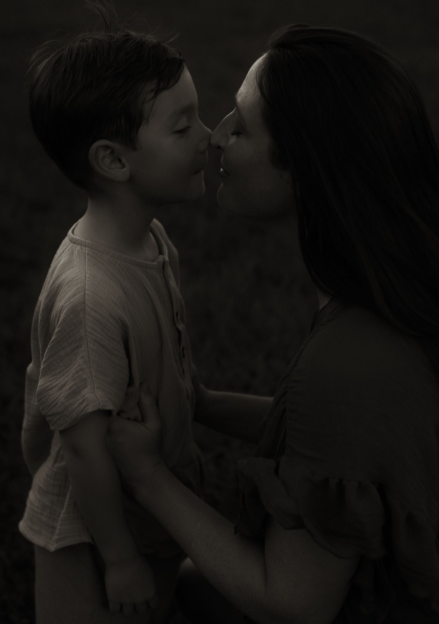 A mother tenderly kisses her young son while holding him in a dark, moody black and white photo.