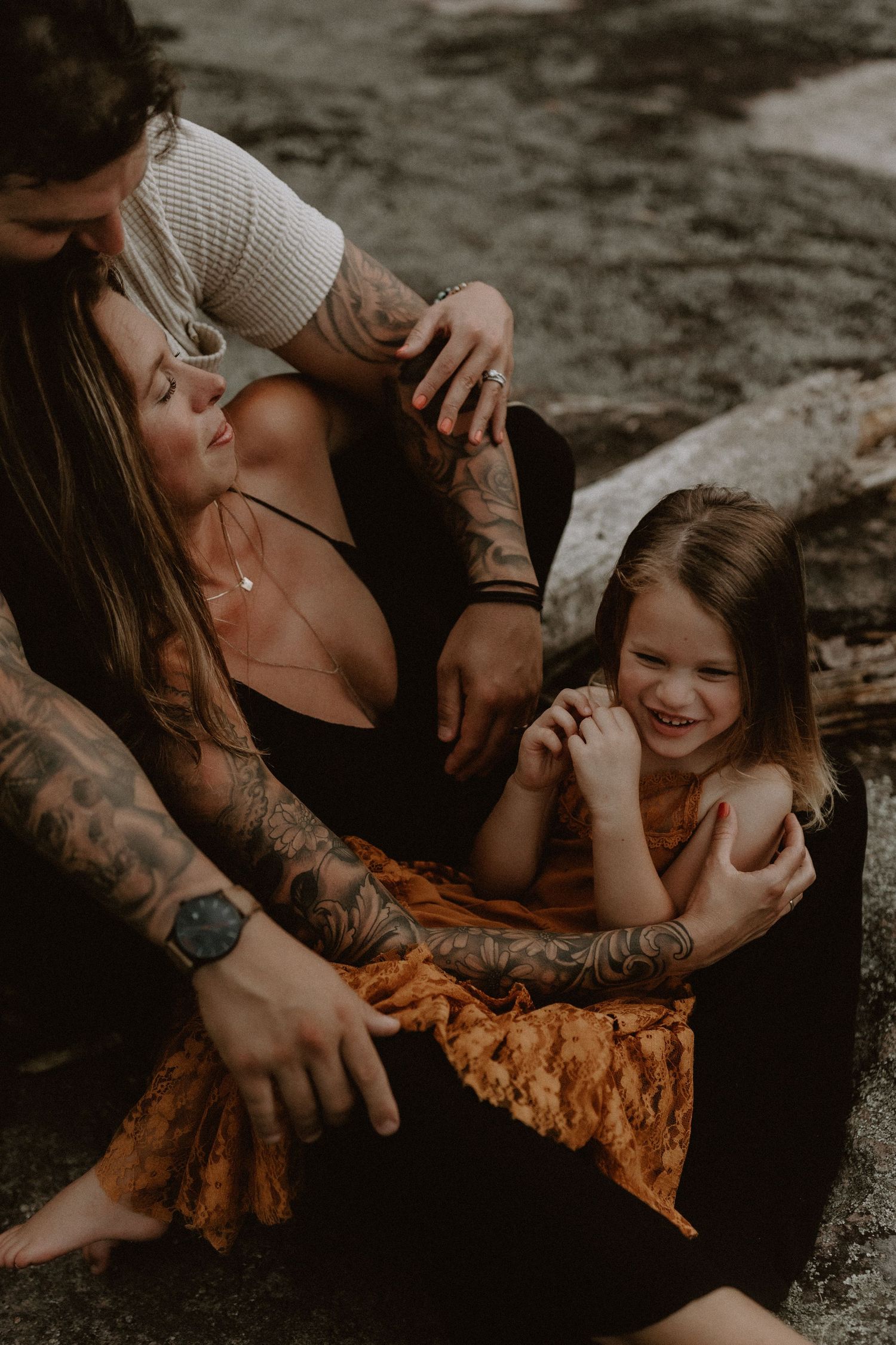 Tattooed couple sitting outdoors with their laughing young daughter in a warm, candid family portrait.
