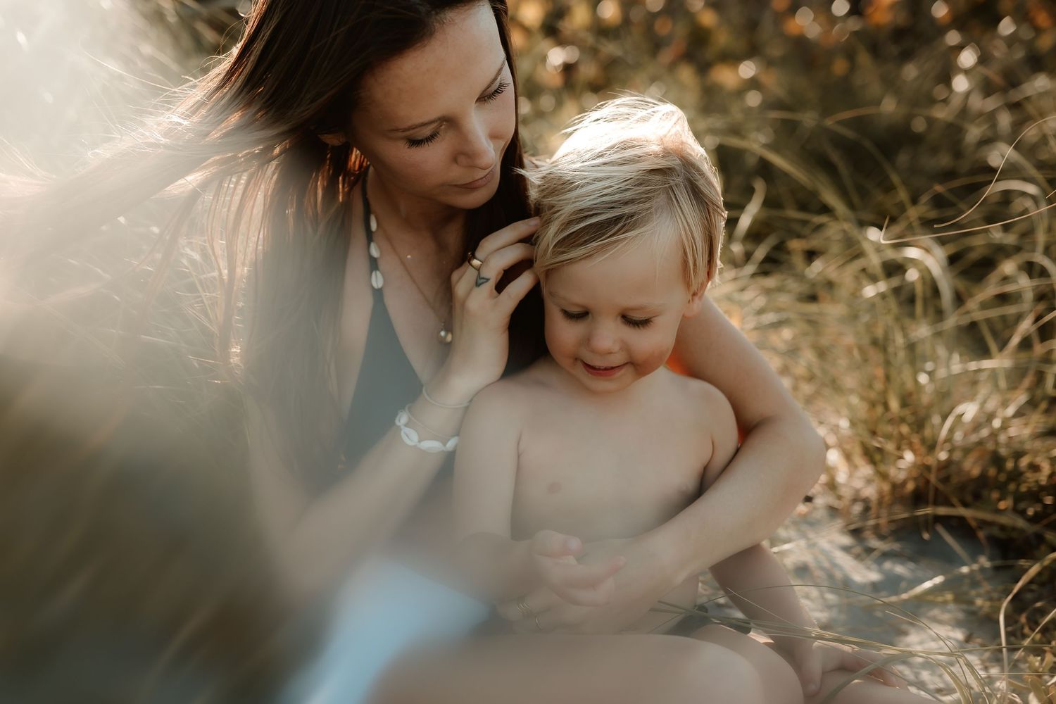 A mother tenderly holds her young blonde toddler outdoors in golden sunlight among tall grass.