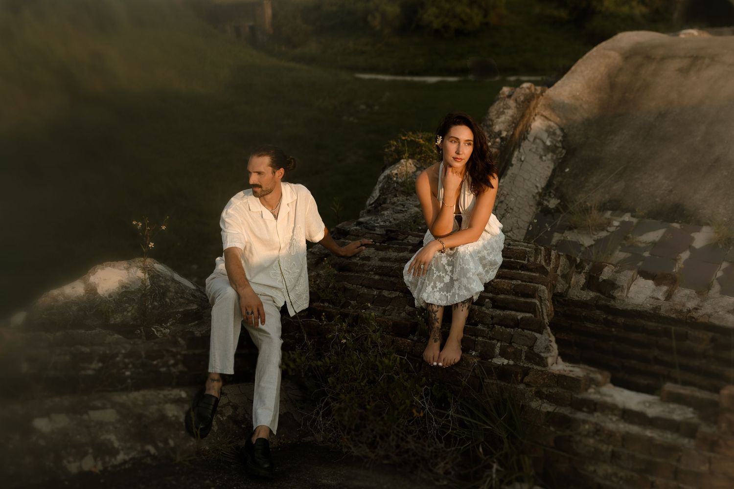 A man and woman sit on ancient stone ruins surrounded by lush greenery in warm golden light.
