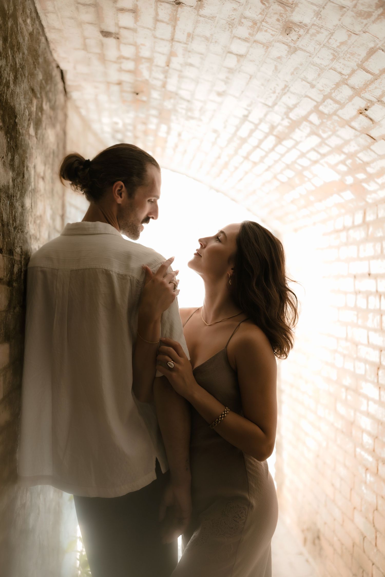 A couple shares a romantic moment in a sunlit brick tunnel, smiling and holding hands in warm, glowing light.