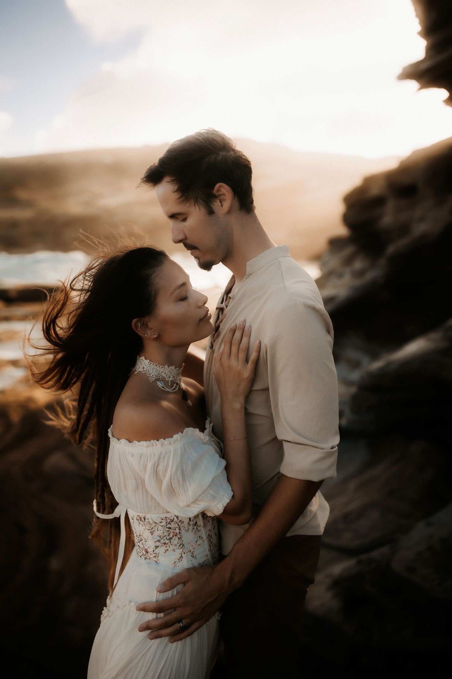 Couple in romantic embrace at golden hour, woman in white boho dress, man in cream shirt, rocky coastal backdrop.