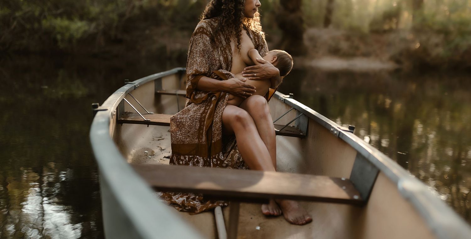 Mother nursing baby while sitting in canoe on peaceful tree-lined river in boho dress.