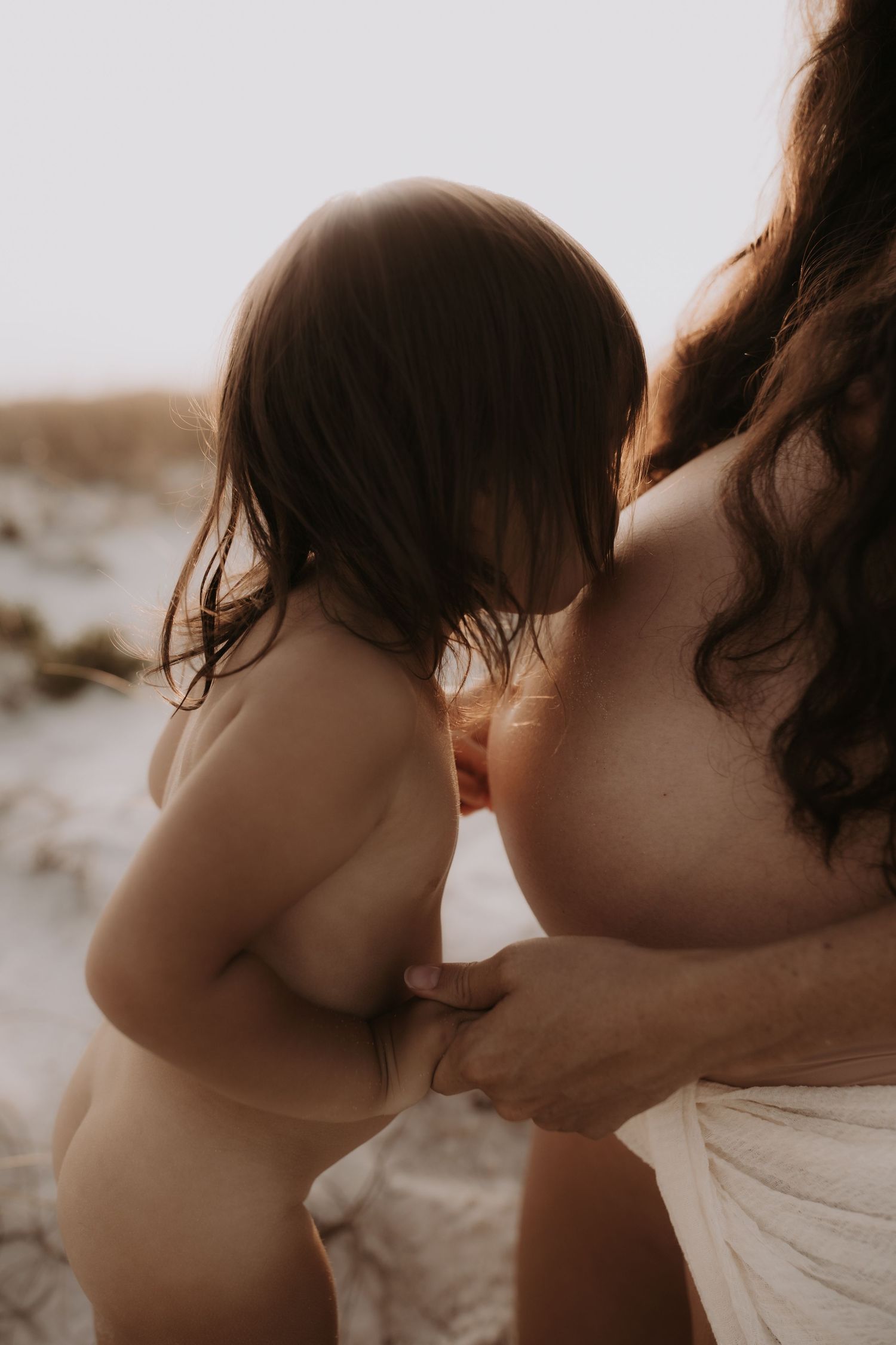 Young toddler with brown hair leans against pregnant mothers belly on sandy beach at sunset.