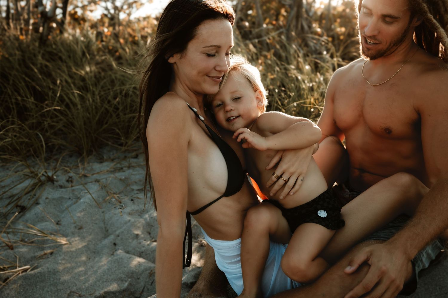 A happy family with a baby enjoys a sunny beach day together, sitting on sand surrounded by golden grass.