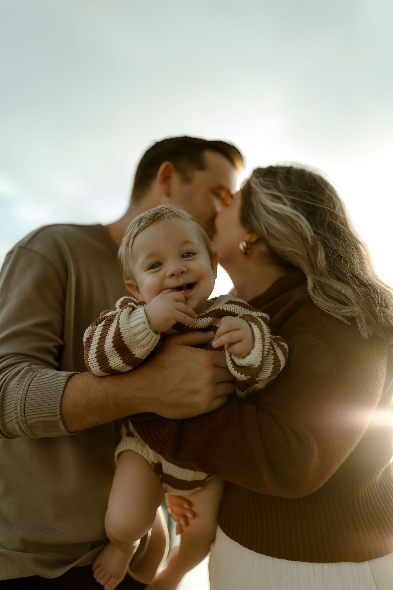 Parents kissing while holding their smiling baby outdoors in warm sunlight.