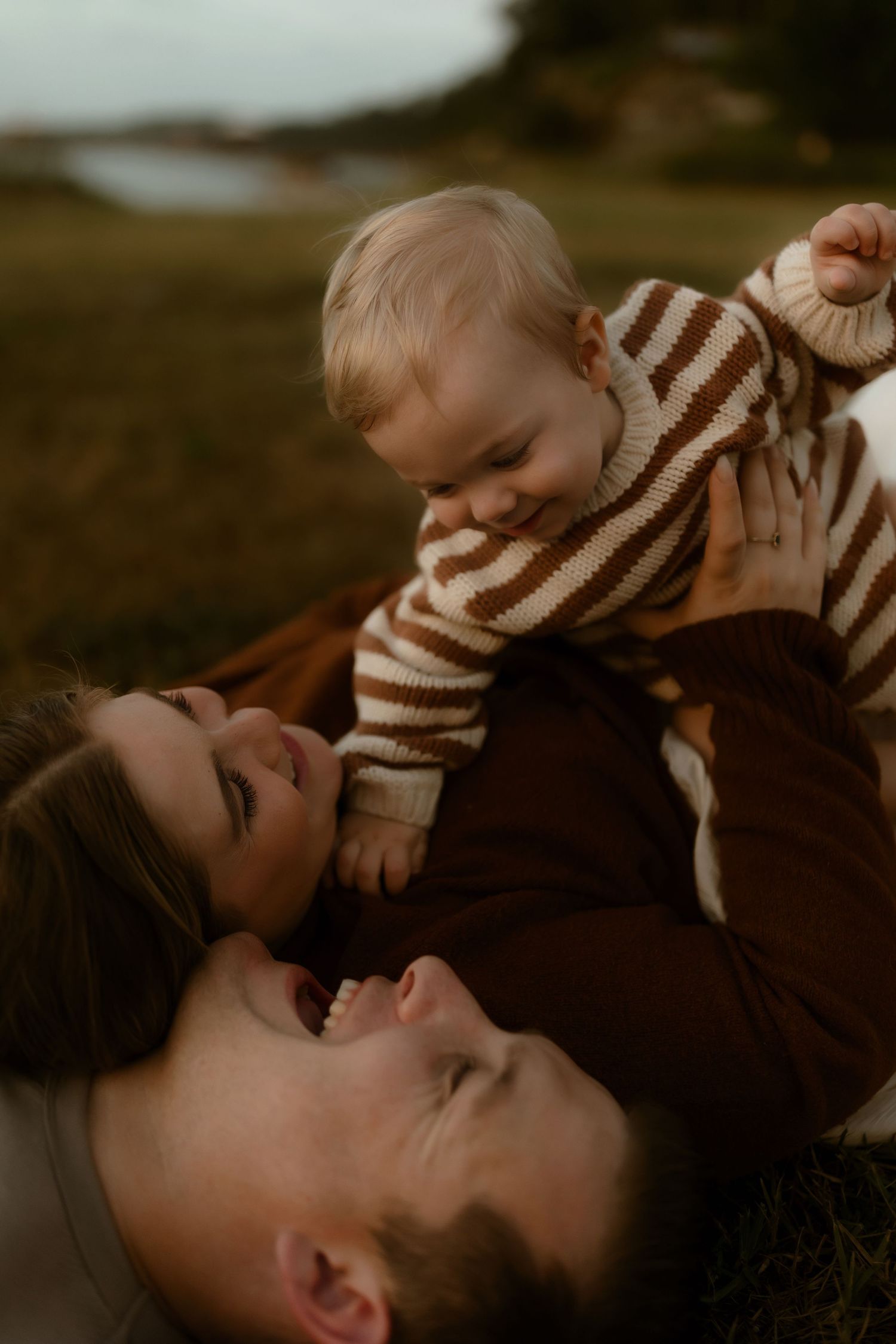 Happy family lying on grass, parents laughing while holding smiling baby in brown striped sweater outdoors.