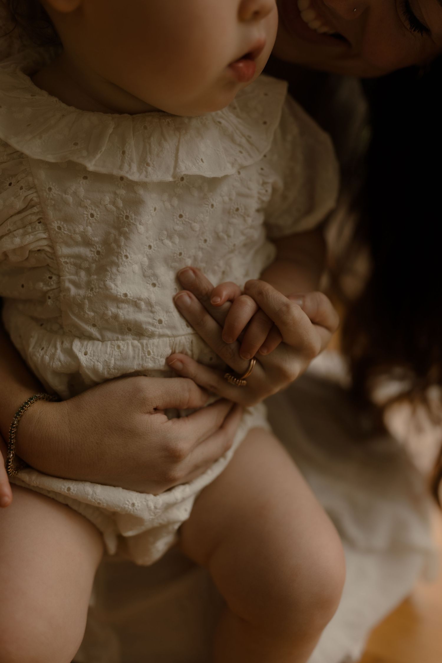 A mother tenderly holds her baby dressed in a white eyelet romper, gently clasping tiny hands together.