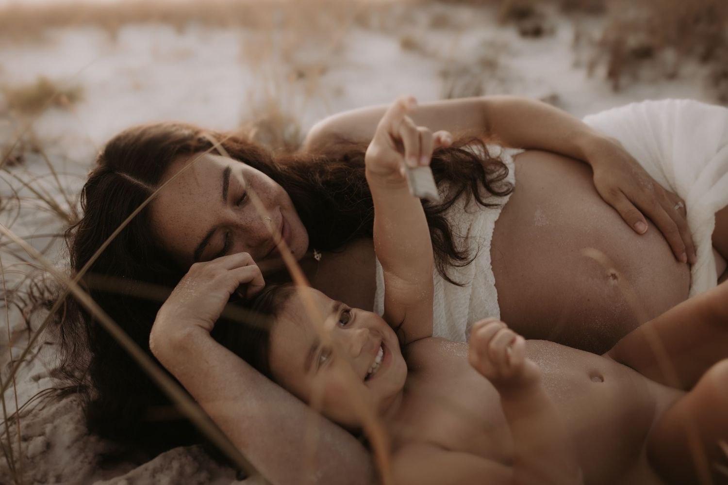 Pregnant woman lying on beach sand with her young child in a warm maternity photography session.