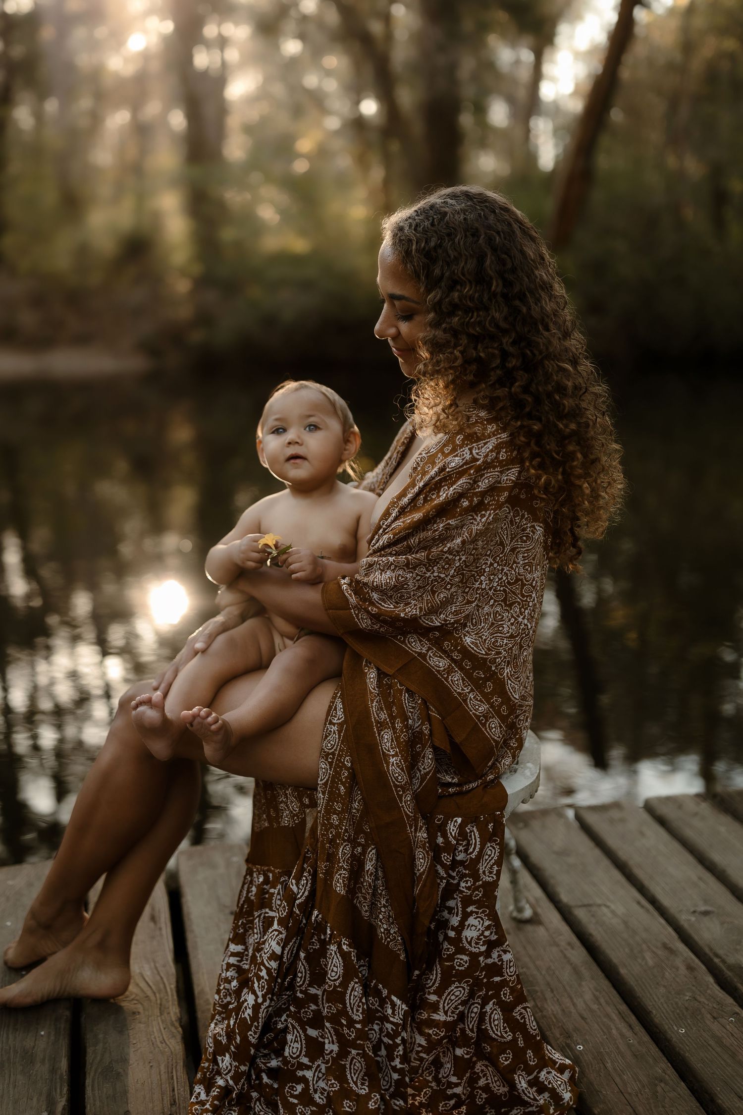 Mother in bohemian dress sits on dock holding baby with yellow flower at sunset.