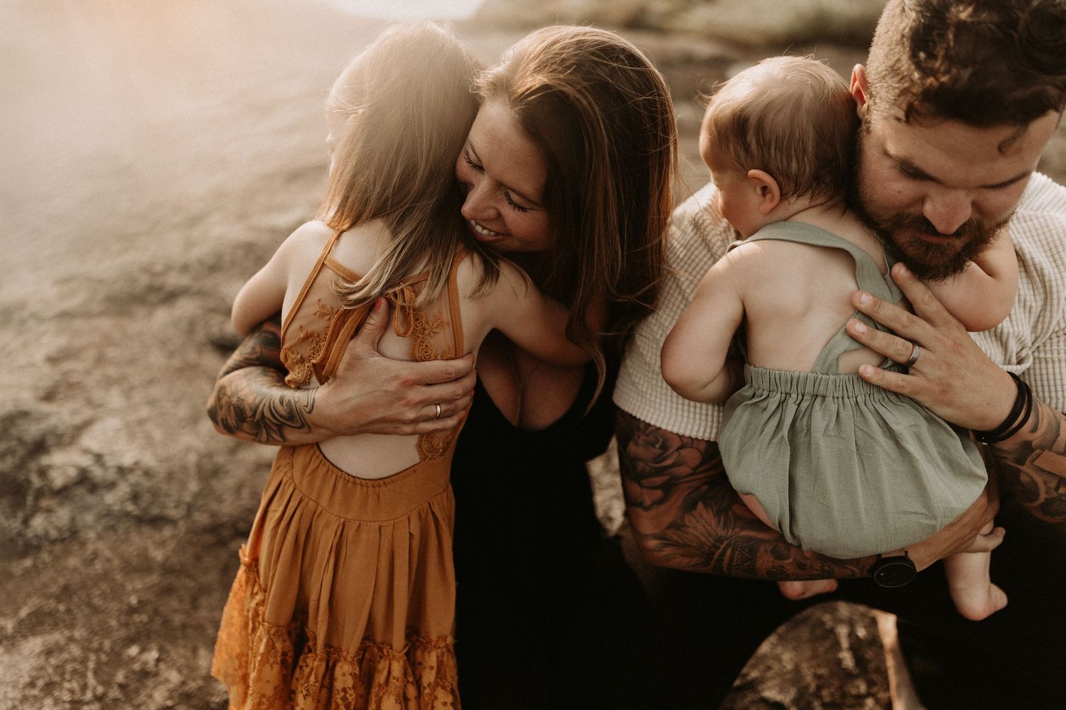 A warm family moment with parents embracing their two young children outdoors in golden sunlight.
