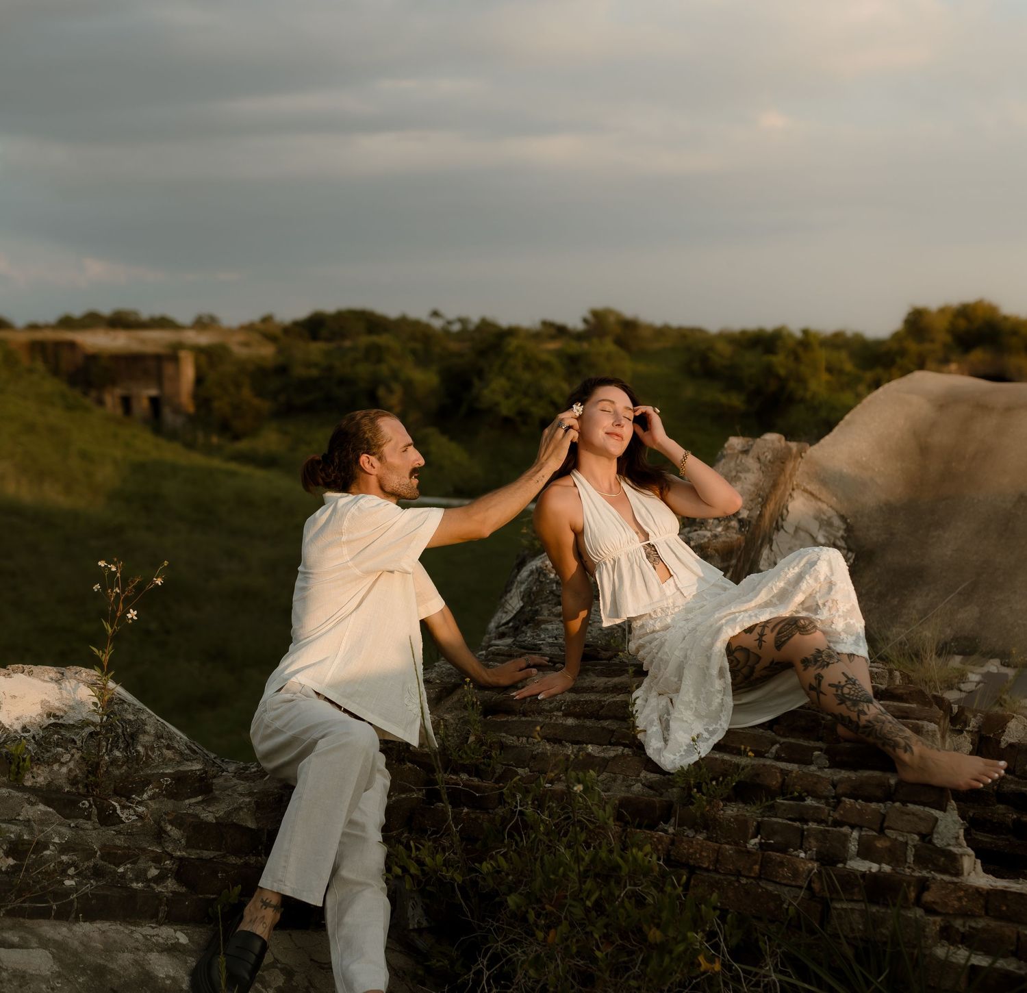 Couple in white outfits laughing and playing on ancient stone ruins at golden hour.