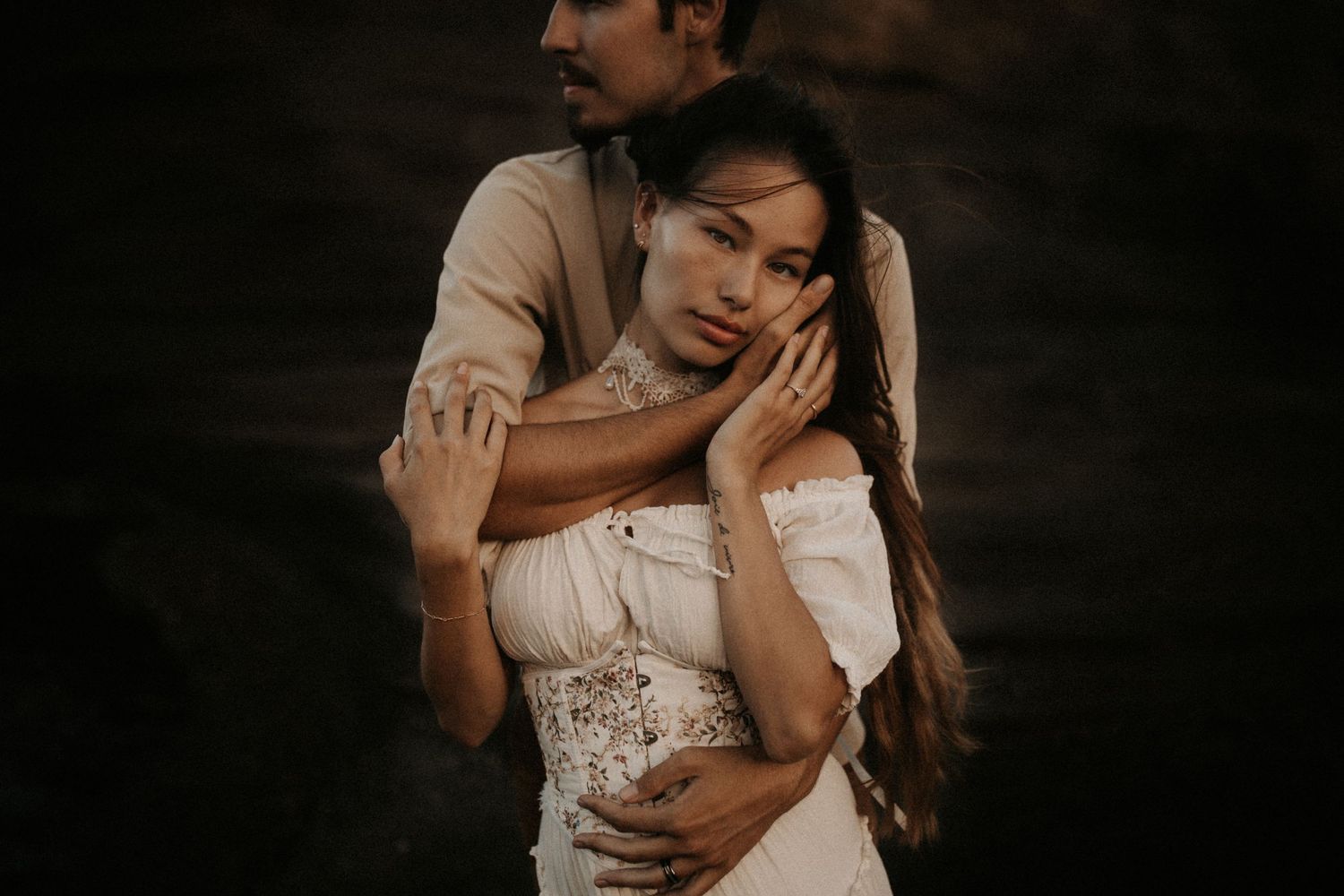 Romantic couple embracing in moody dark setting, woman in white off-shoulder dress with long hair, man holding her tenderly.