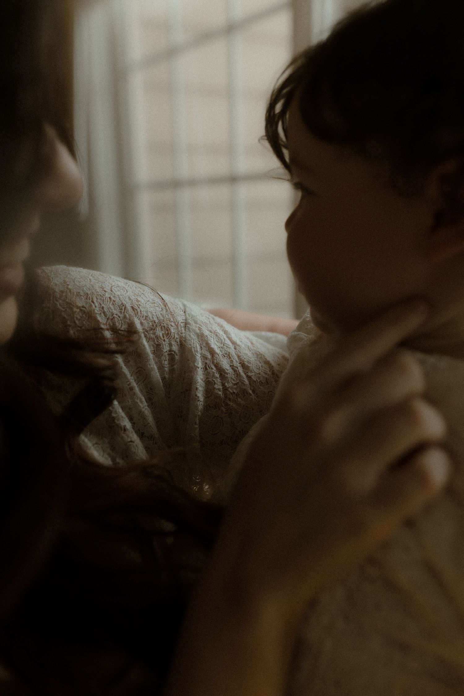 A mother tenderly holds her newborn baby near a window in soft, moody light.