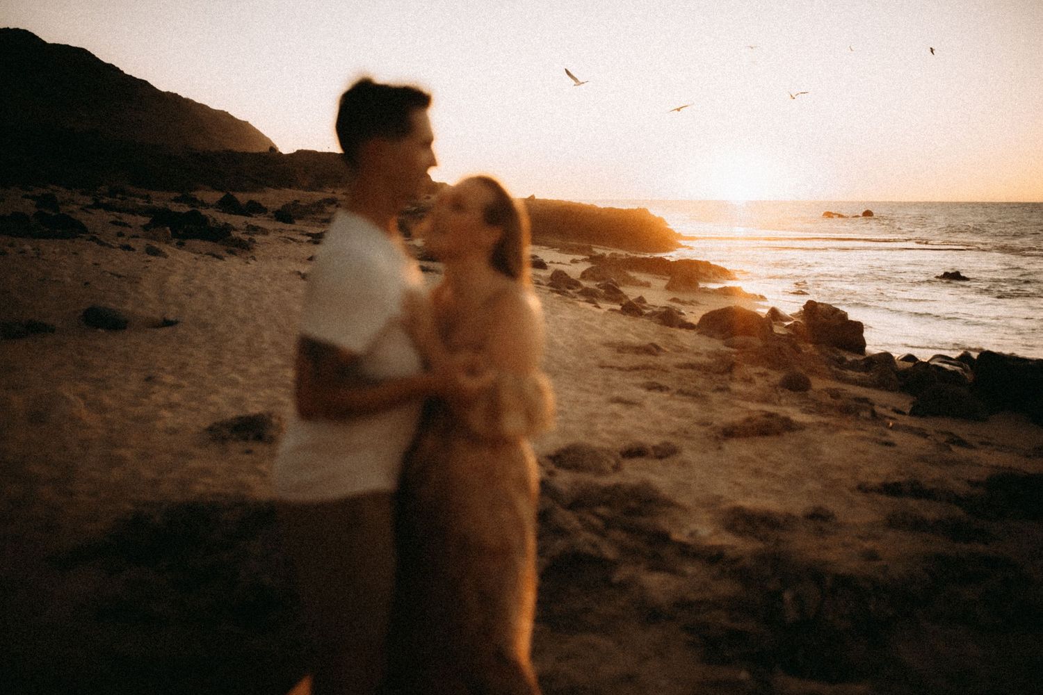 Romantic couple embracing on rocky beach at golden sunset with ocean waves in background.