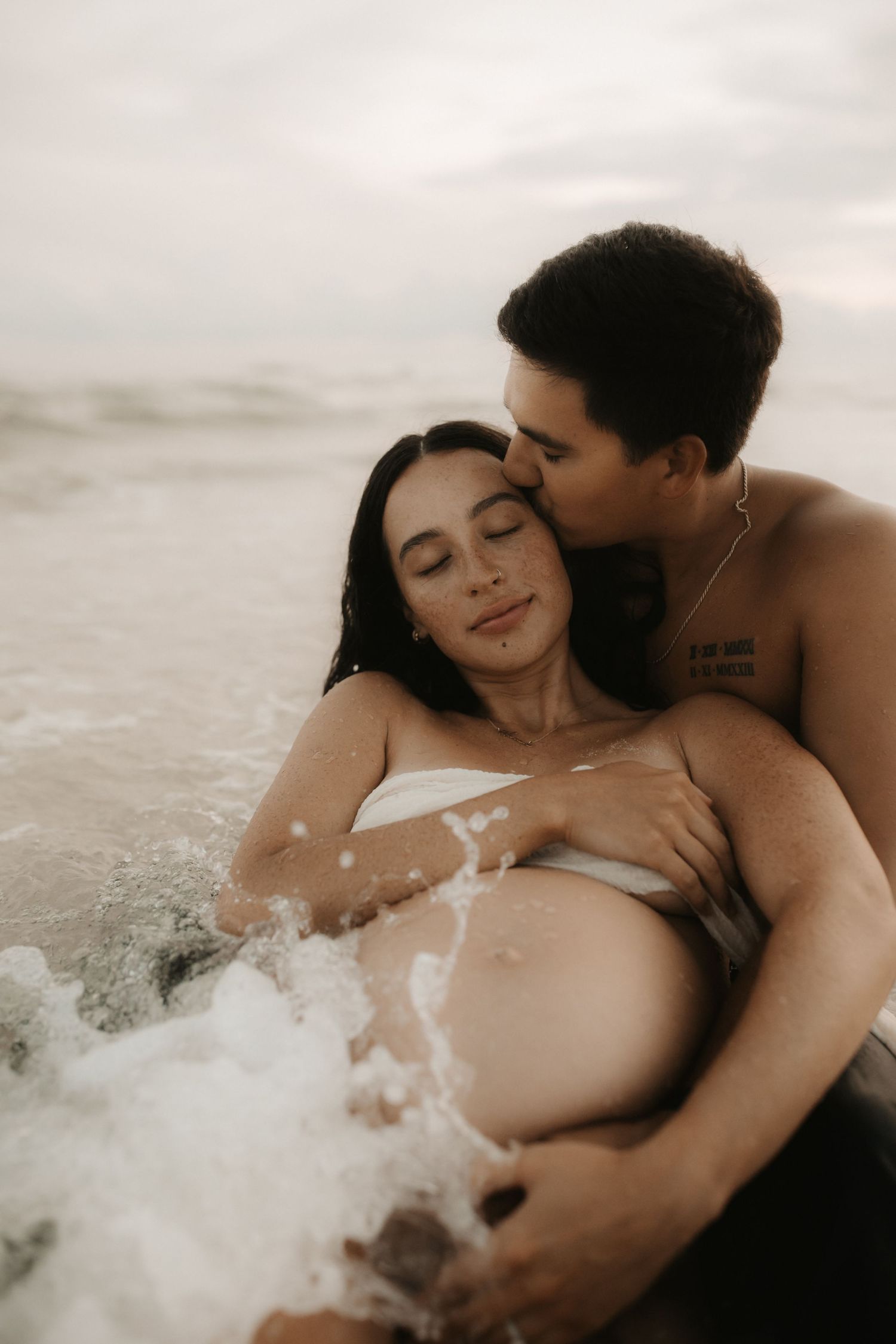 Couple sharing tender beach moment, man kissing pregnant womans cheek while ocean waves splash around them.