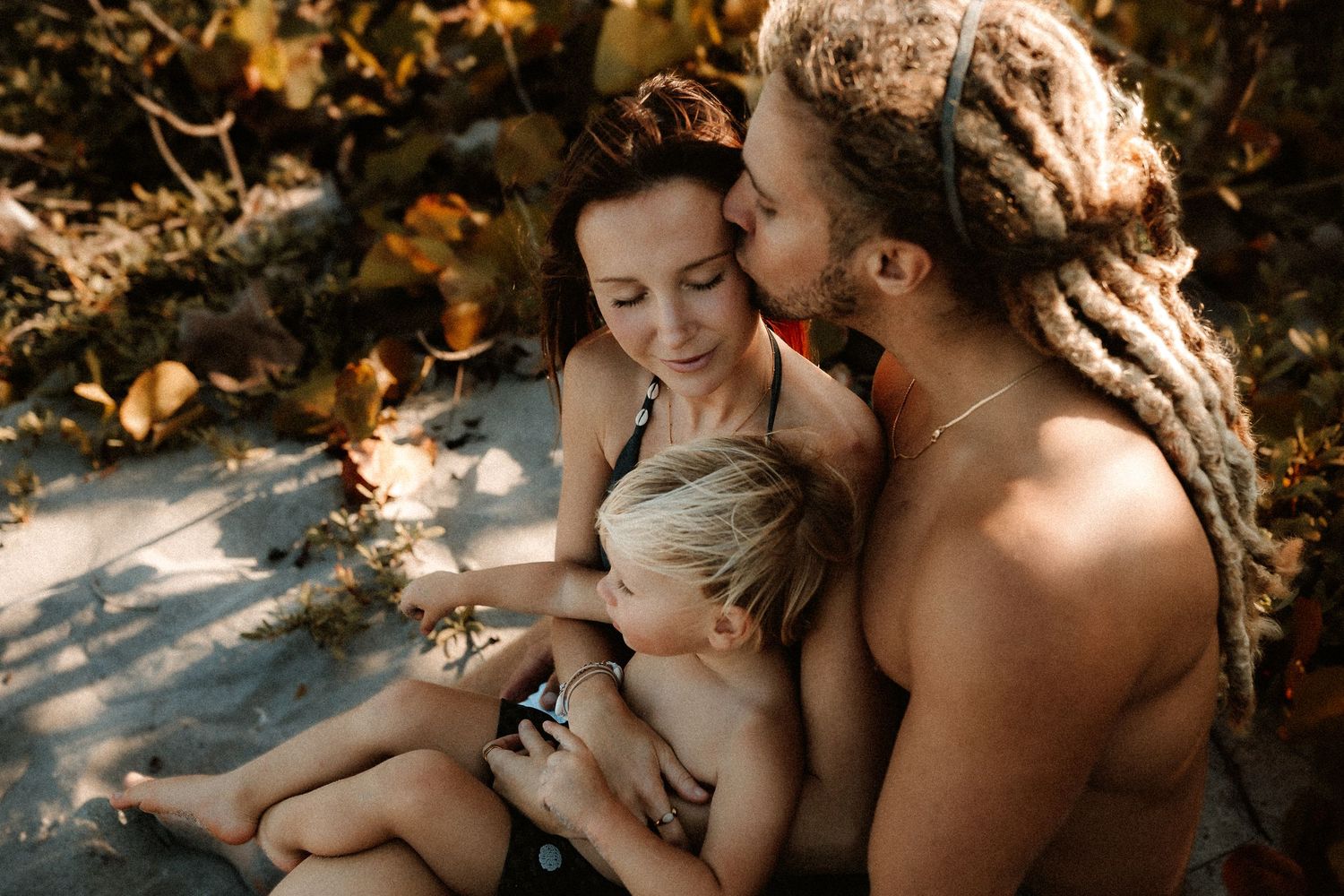 Family of three sitting on sandy beach, parents embracing blonde toddler outdoors.