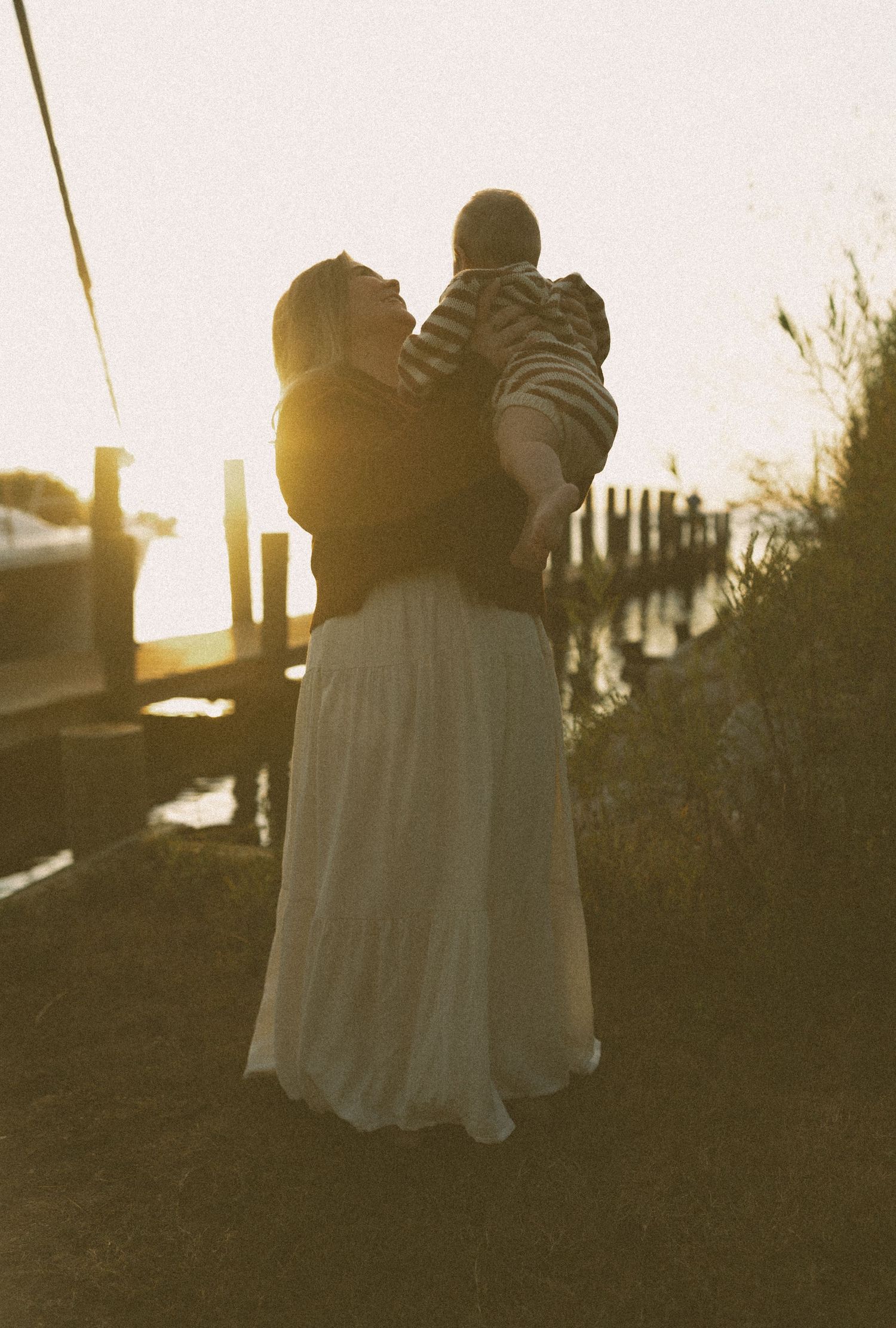 Mother lifting baby at golden hour near a waterfront dock at sunset.