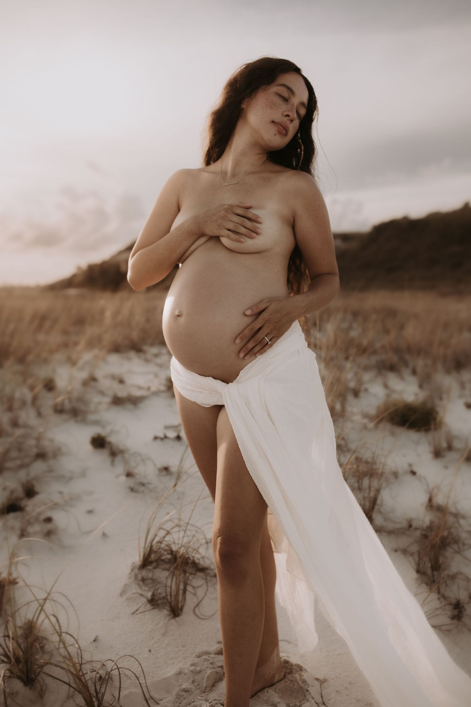 Pregnant woman in white fabric wrap posing on sandy beach dunes at golden hour sunset.