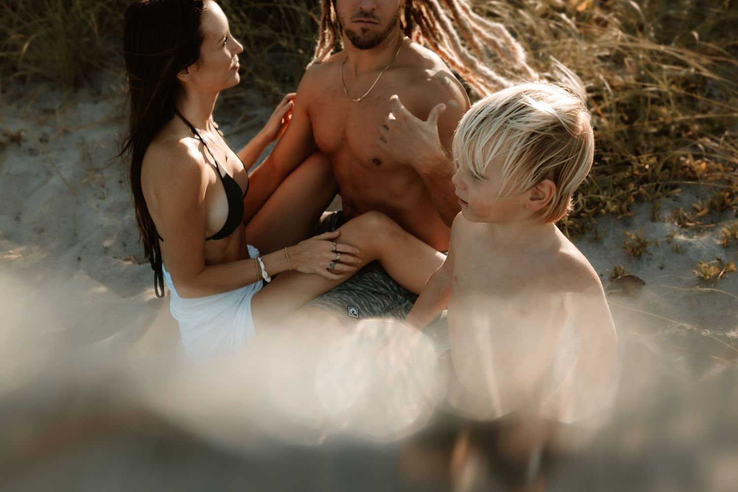 Family sitting together on sandy beach dunes, parents and child enjoying a sunny day outdoors.