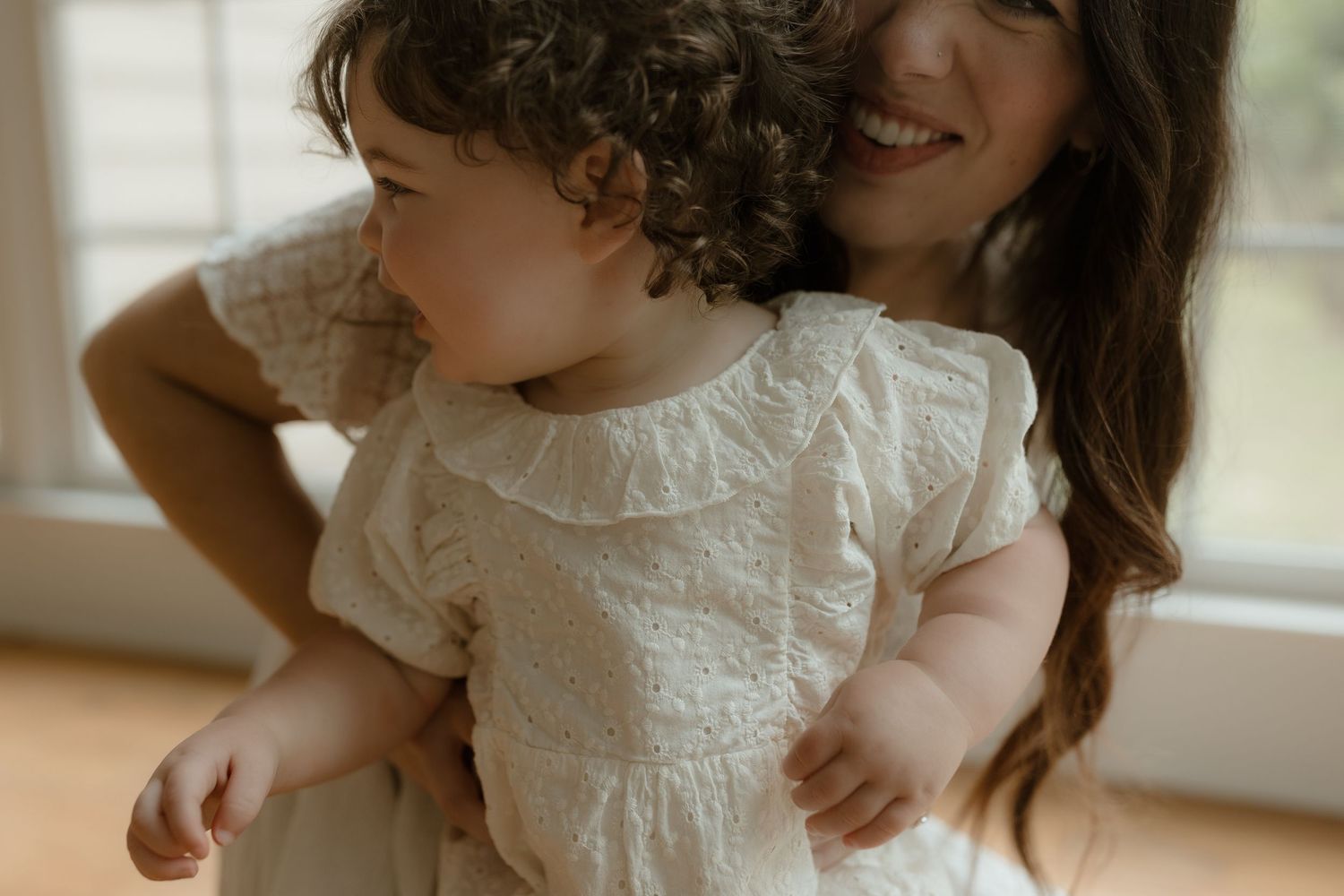 A smiling mother in a white dress holds her curly-haired toddler, both wearing matching white outfits indoors.