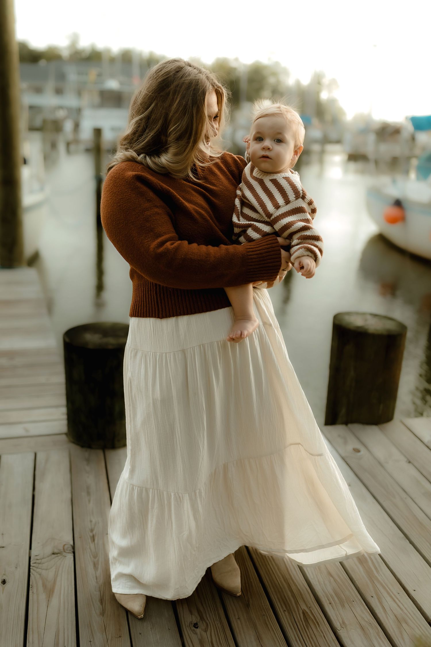 Woman in brown sweater and white maxi skirt holding a baby on a wooden dock near a marina.