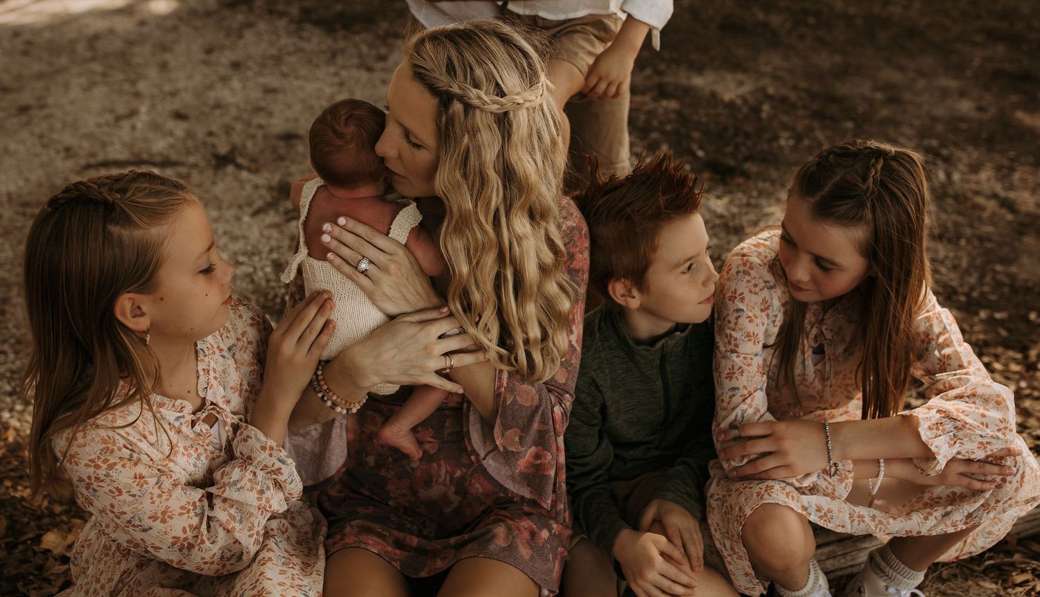 A mother sits outdoors holding a newborn baby surrounded by her children in floral outfits in a warm family portrait.