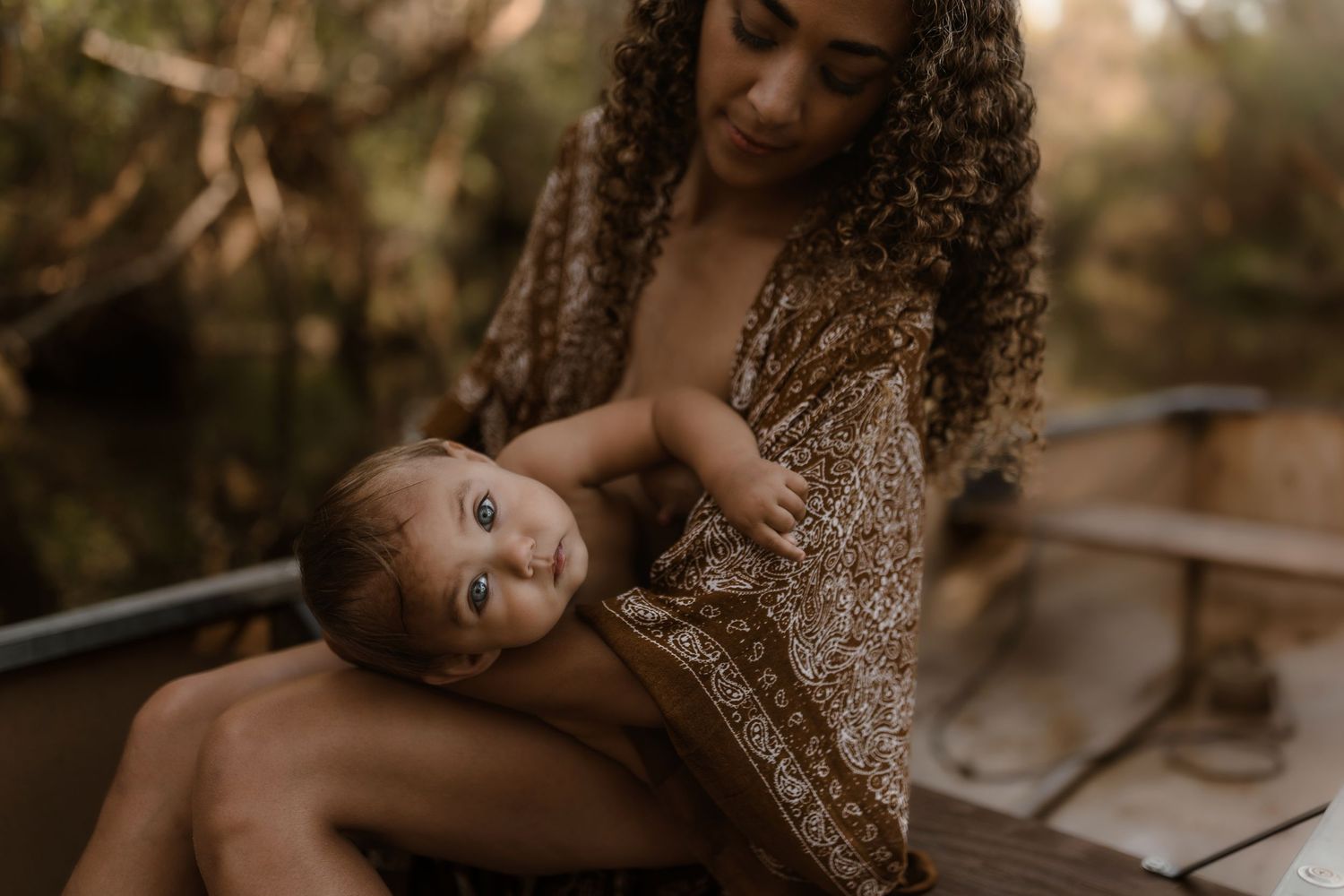 A mother cradles her wide-eyed baby in her arms while sitting in a boat in a natural setting.